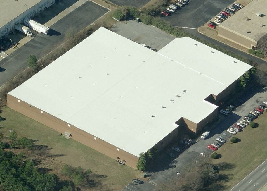 Aerial view of a large commercial building with a white flat roof, surrounded by parking lots with cars and a treeline.
