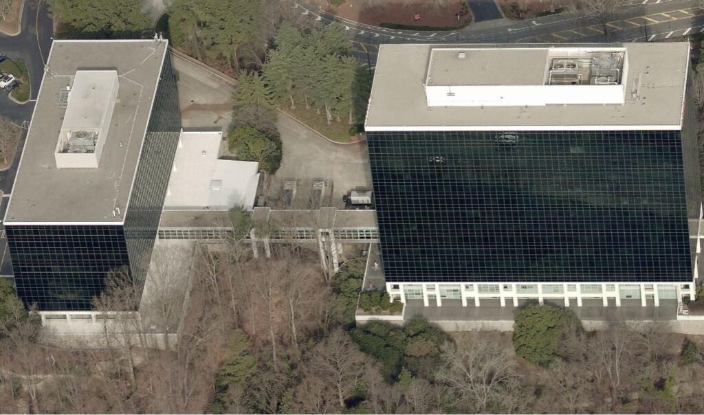 Aerial view of two glass-paneled office buildings connected by a covered walkway over a wooded area.