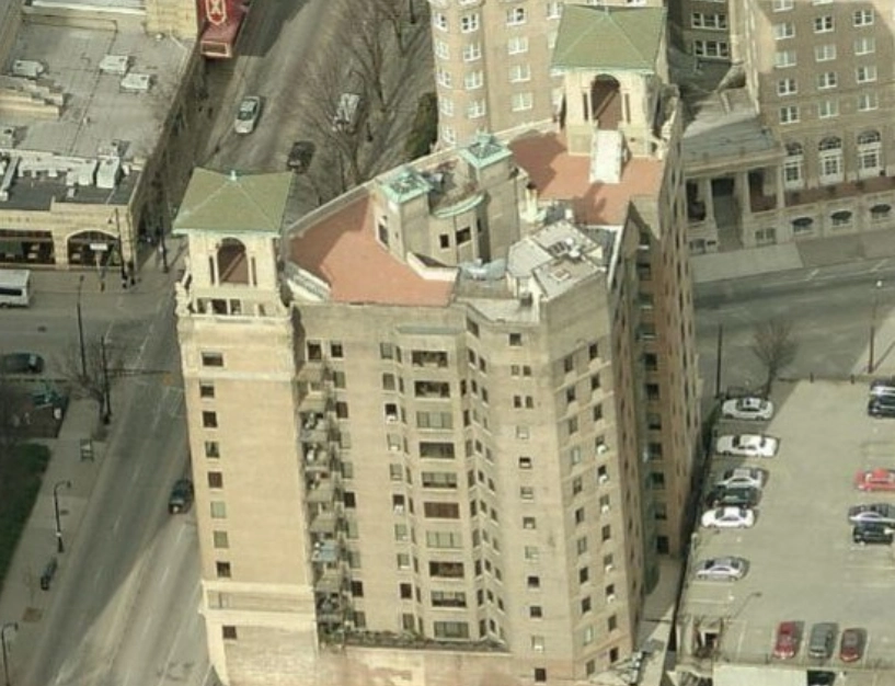 Aerial view of a tall, beige, multi-story building with a flat roof and corner towers, situated between city streets and a parking lot.