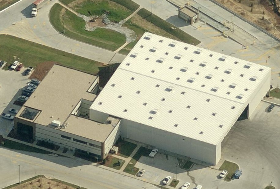 Aerial view of a large commercial building featuring a warehouse with a metal roof and an attached office section, surrounded by parking lots and access roads.