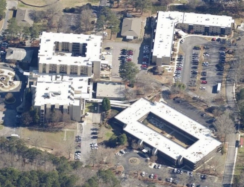 Aerial view of three large commercial buildings with flat white roofs, surrounded by parking lots and wooded areas.
