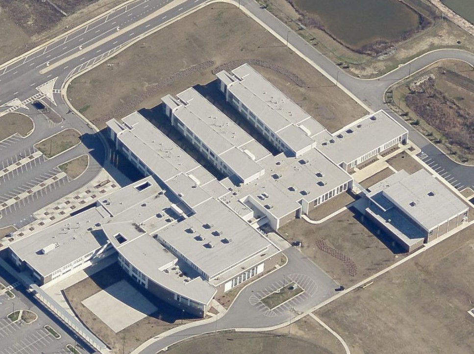 Aerial view of a large school building complex with multiple interconnected wings, flat roofs, and surrounding parking lots.