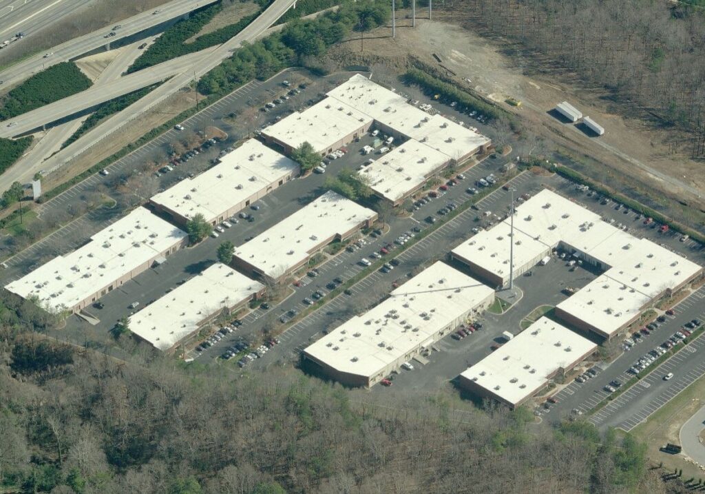 An aerial view of an office park complex featuring several flat-roofed buildings surrounded by parking lots and wooded areas, located next to a highway interchange.