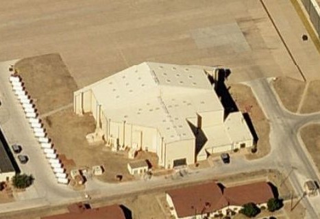 Aerial view of a large, angular industrial hangar building next to a row of parked trailers.