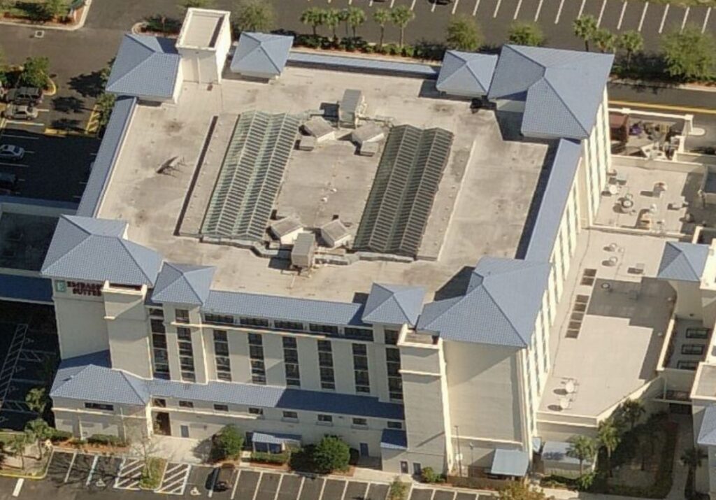 Aerial view of a multi-story hotel with a blue-tiled roof, two large rectangular skylights, and a parking lot.