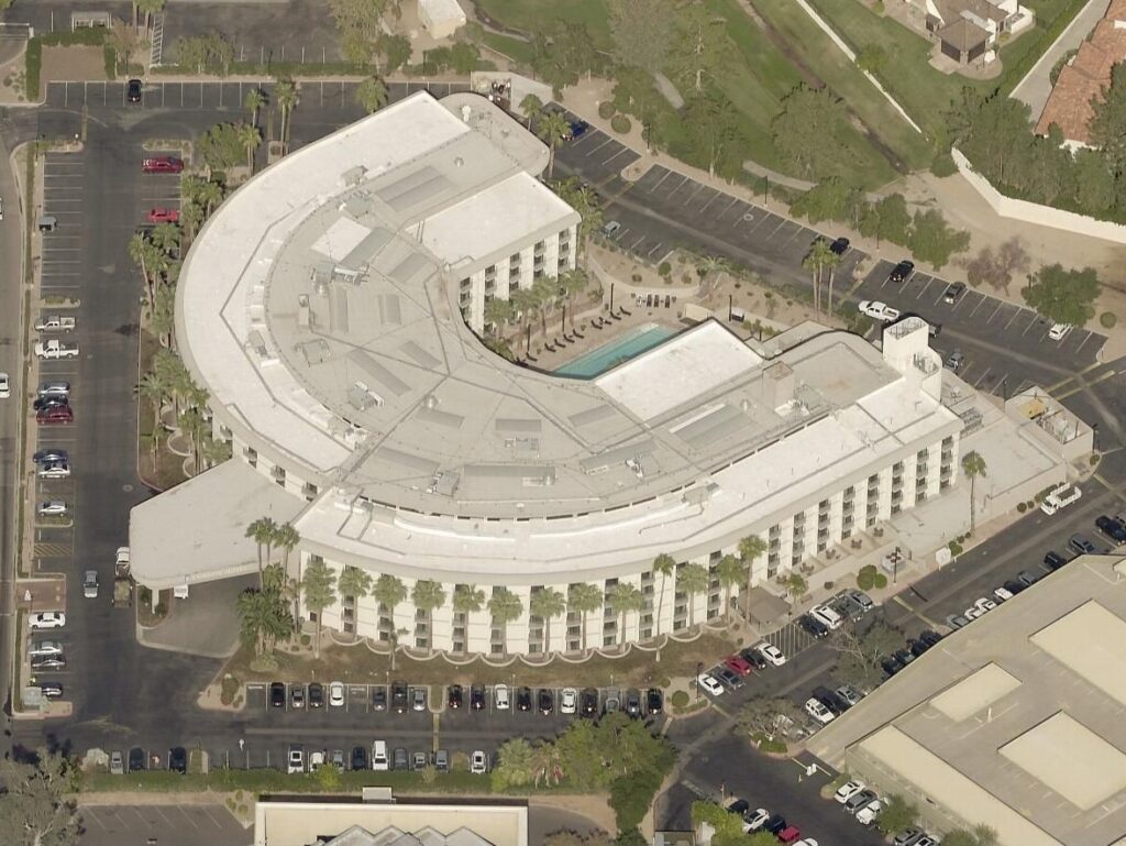 An aerial view of a crescent-shaped hotel building with an outdoor pool, surrounding parking lots, and palm trees.