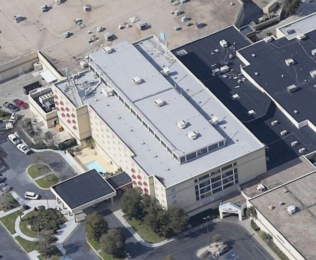 An aerial view of a multi-story hotel building with a flat roof, an outdoor swimming pool, and an attached parking lot.