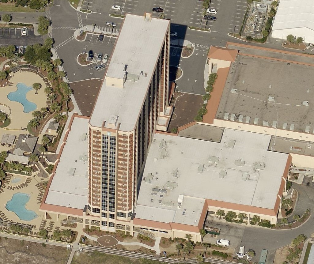 Aerial view of a tall hotel tower connected to a lower conference building, with nearby swimming pools and parking lots.