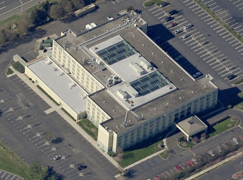 Aerial view of a multi-story hotel building with a flat roof featuring skylights, surrounded by parking lots.