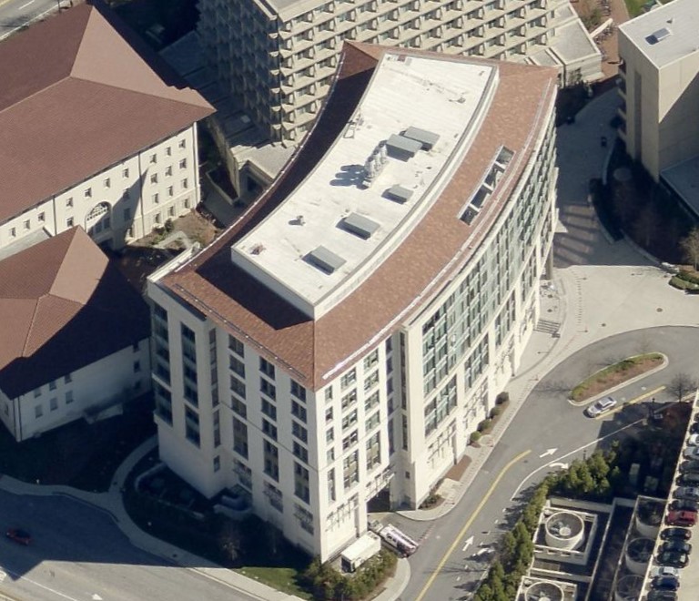 Aerial view of a large, curved office building with a brown roof, situated among surrounding campus buildings and parking areas.