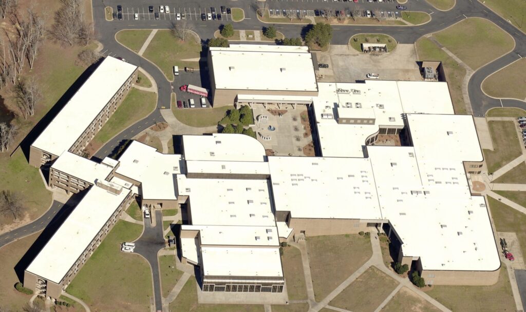 Aerial view of a large school or office complex with a tan exterior and white flat roofs, surrounded by parking lots and grassy areas.