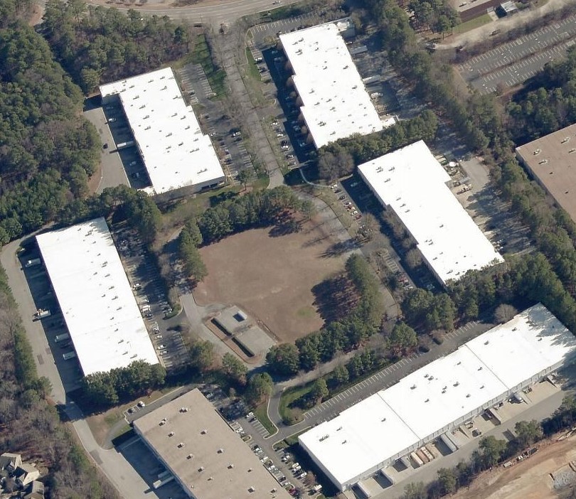 Aerial view of an office park featuring six warehouse-style buildings with white roofs surrounding a central lawn and parking lots.