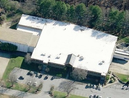 Aerial view of a large commercial building with a white roof, surrounding parking lots, and trees.
