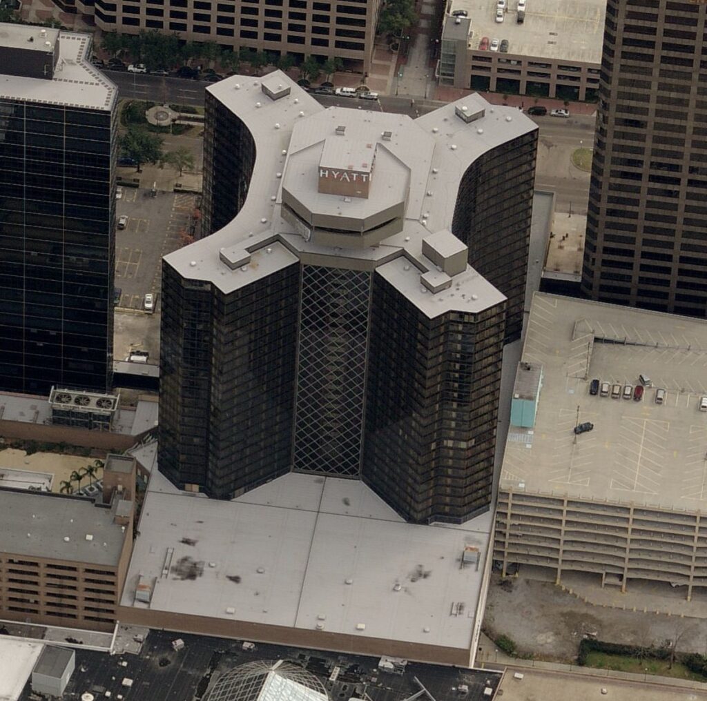 Aerial view of a cross-shaped Hyatt hotel building surrounded by taller skyscrapers and parking structures.