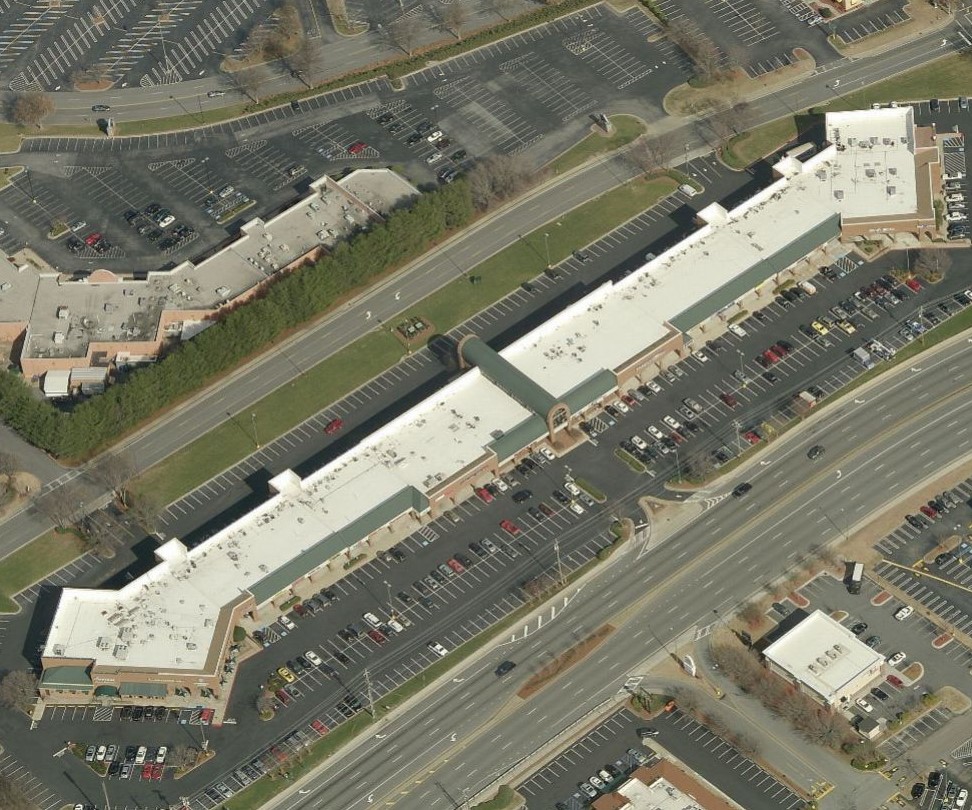 Aerial view of a long, single-story commercial strip mall building with large parking lots, surrounded by roads and other nearby parking areas.