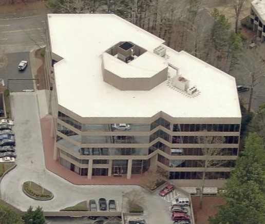An aerial view of a multi-story office building with an irregular footprint, featuring glass-paneled walls and a flat roof with mechanical units.