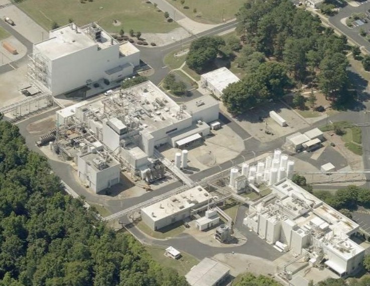Aerial view of an industrial manufacturing complex with multiple connected white buildings, outdoor storage tanks, and surrounding trees.