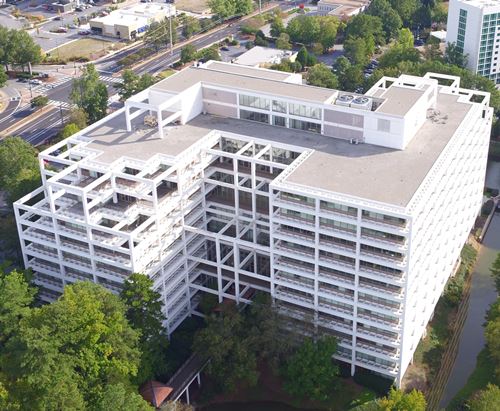 Aerial view of a large white office building with a complex structural frame design surrounded by trees and roads.