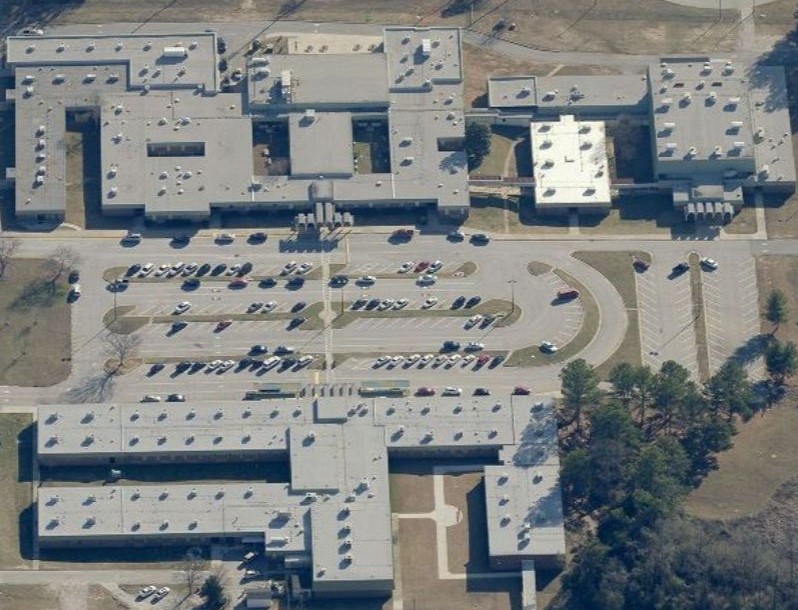 Aerial view of a school campus featuring two large buildings, parking lots with cars, and surrounding trees.