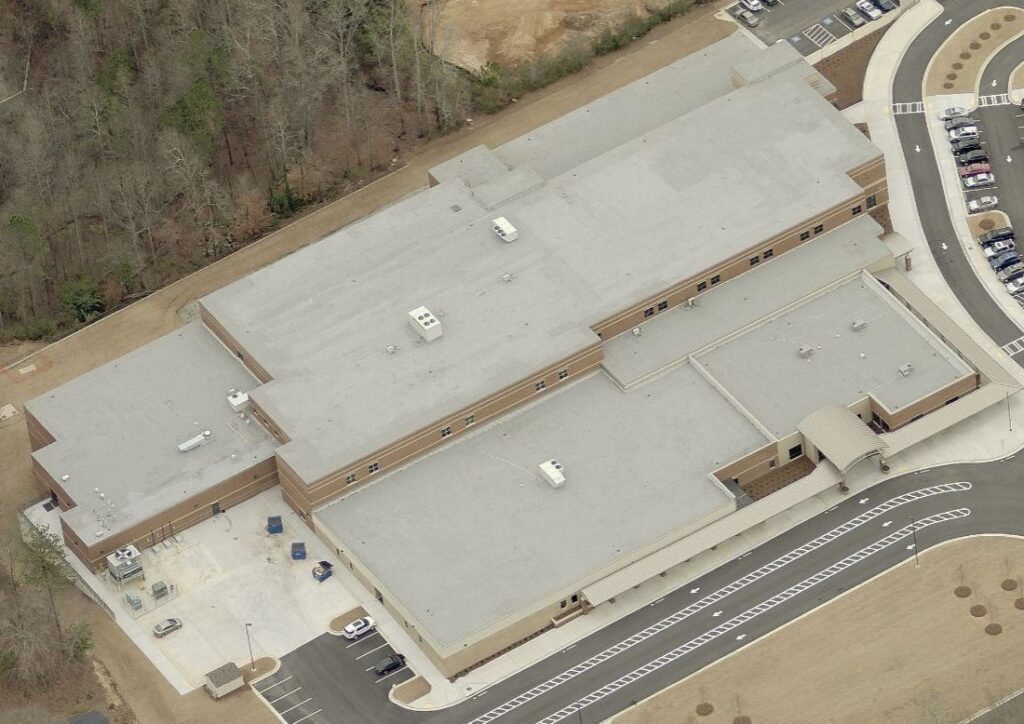 Aerial view of a large rectangular school building with a flat roof, surrounded by parking lots and wooded areas.