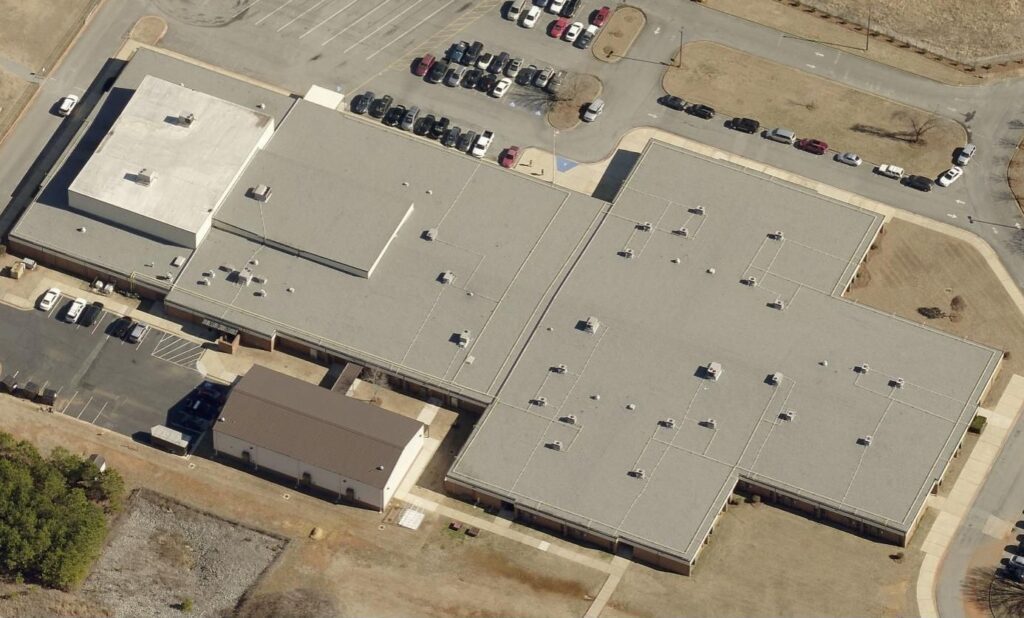 Aerial view of a large school building with a gray roof and adjacent parking lots filled with cars.