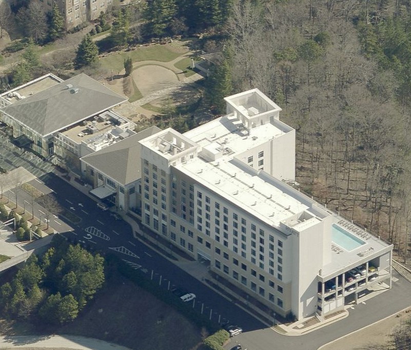 Aerial view of a multi-story hotel with an attached parking deck, a rooftop pool, and surrounding wooded grounds.