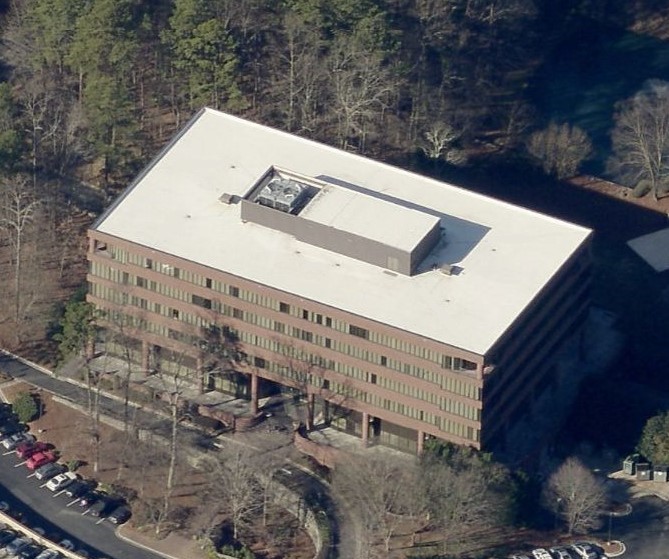 An aerial view of a multi-story, rectangular brick office building situated in a wooded area with an adjacent parking lot.