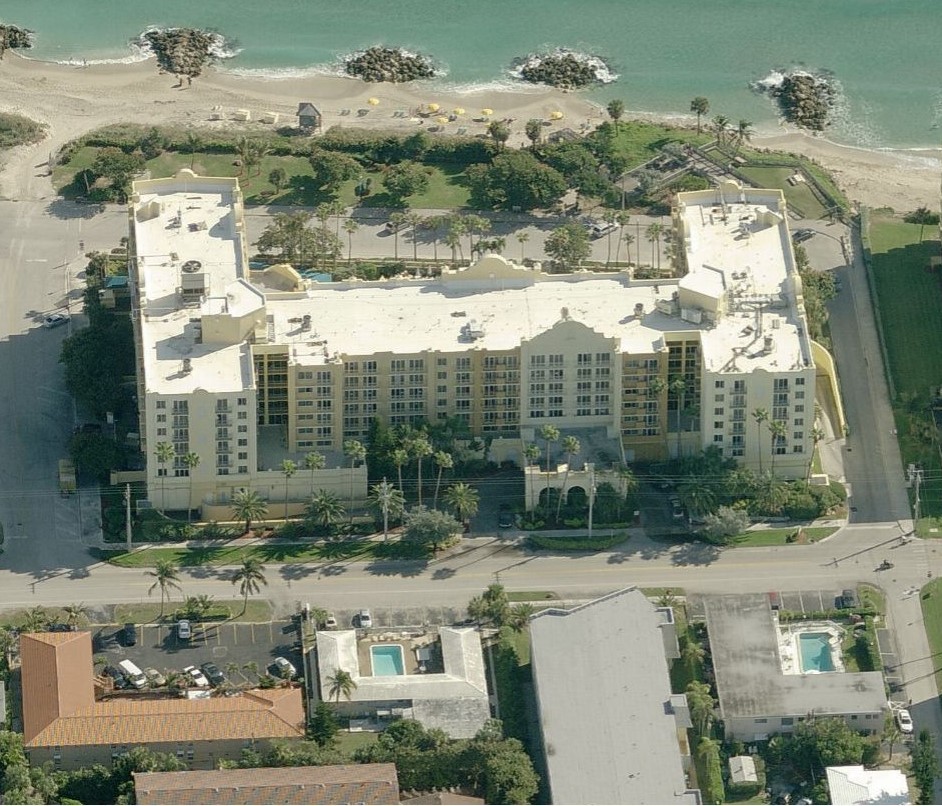 Aerial view of a U-shaped resort hotel situated on a beach with surrounding palm trees and parking areas.