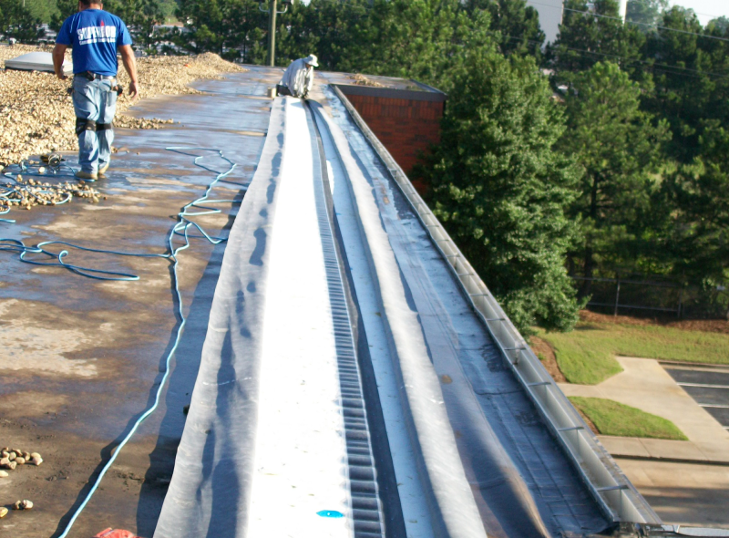 Two roofers install a long strip of roofing membrane on a gravel-covered flat roof.